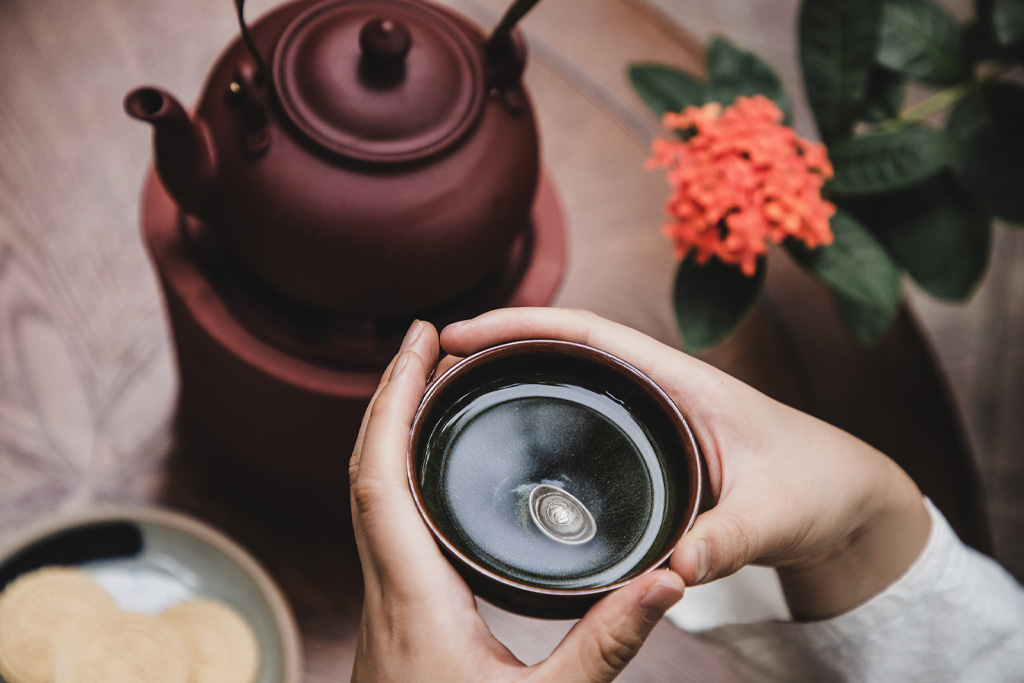 Egy női kéz, amint egy csésze teát tart a kezében, a háttérben teáskanna és keksz látható