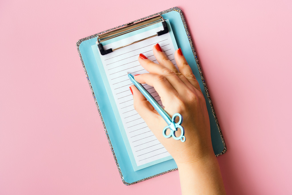 Rózsaszín alapon egy jegyzetfüzet látható, és egy női kéz, amint egy tollat fog