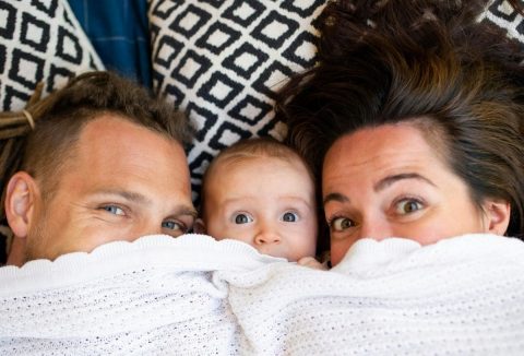 Család fehér takaró mögé bújva, balra apuka, középen kisbaba, jobbra anyuka, mögöttük fekete-fehér mintás díszpárnák