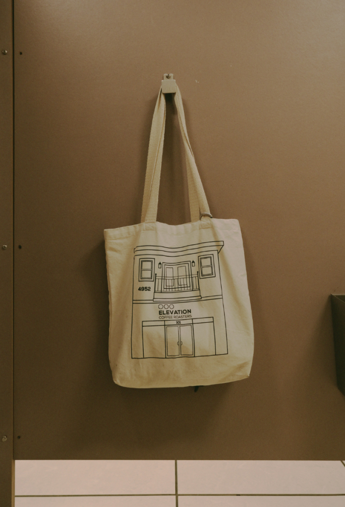 Vászontáska stilizált épület rajzával akasztón lóg barna fal előtt minimalista fotón