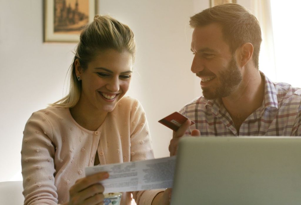 penzugyek-sporolas-milliomos | Marie Claire Fiatal férfi és nő számítógép előtt bankkártyát és értékpapírt tart a kezében, mosolyogva néznek egymásra