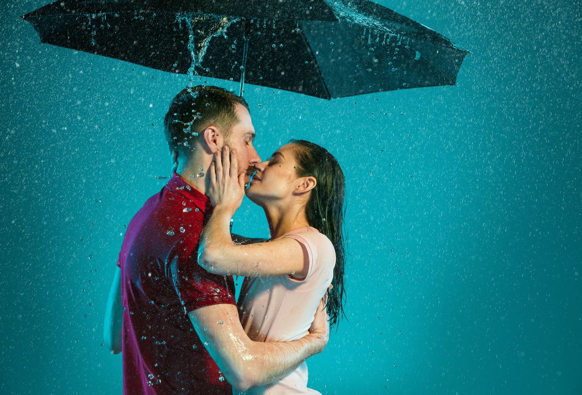 Élénk kék háttér előtt esőben, fekete esernyő alatt csókolózó pár, a férfi piros, a nő fehér pólóban