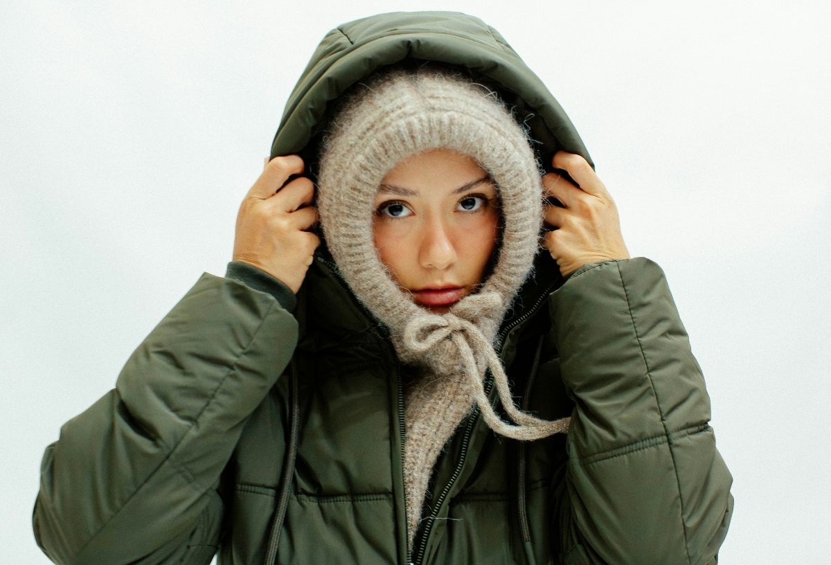 Fiatal nő natúr színű kötött sálsapkában és zöld pufikabátban a fejére húzza a kapucnit