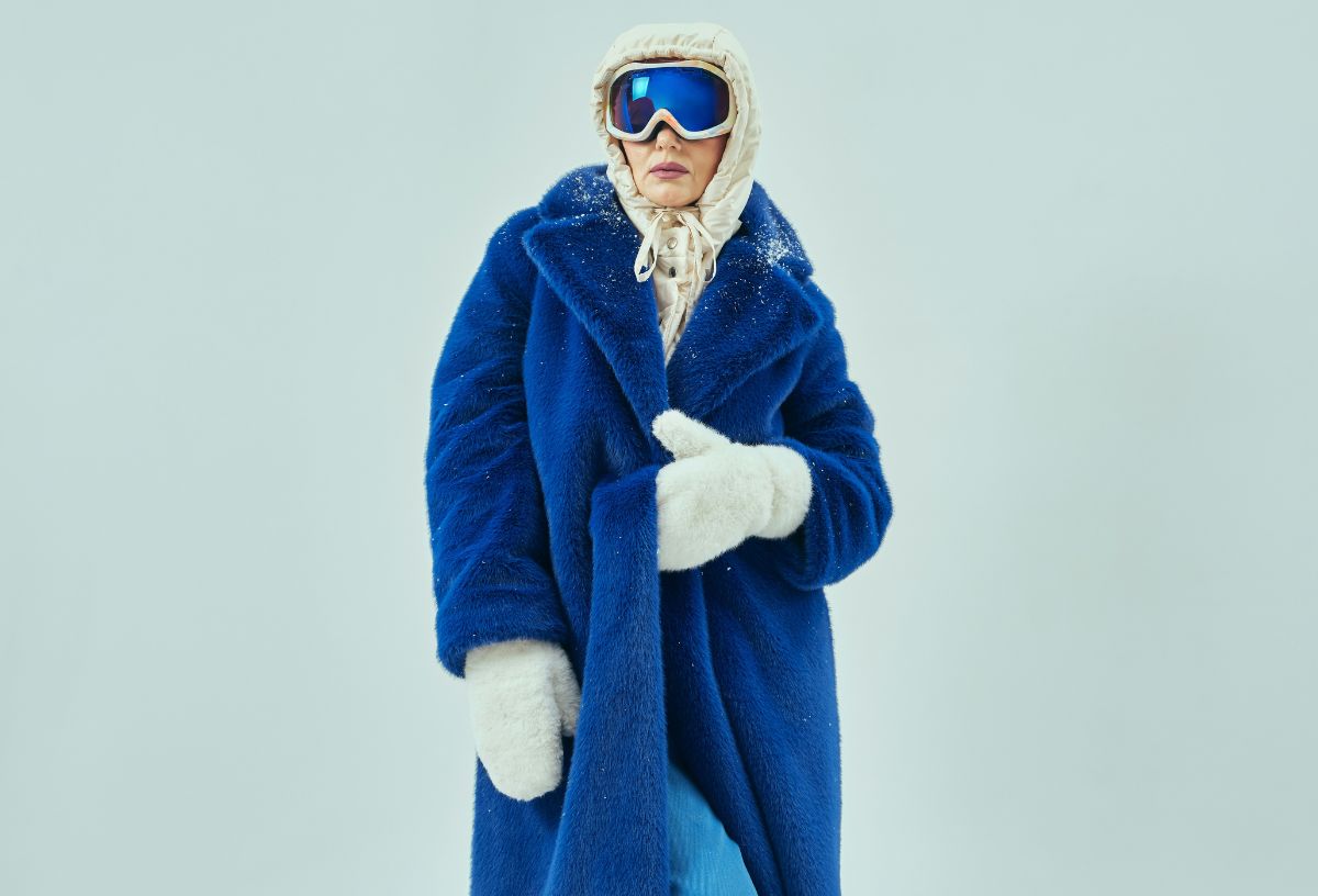 Nő kék műszőrme hosszú télikabátban, fehér egyujjas kesztyűben és fehér kapucniban, kék síszemüveget visel semleges világoskék háttér előtt
