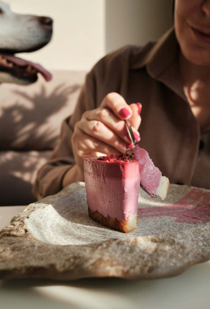 Nő közeli képen egyedi tényérról piros gyümölcsös joghurttortát eszik, egy falatot épp a villlájára szúr, a háttérben elmosódva lihegő kutya
