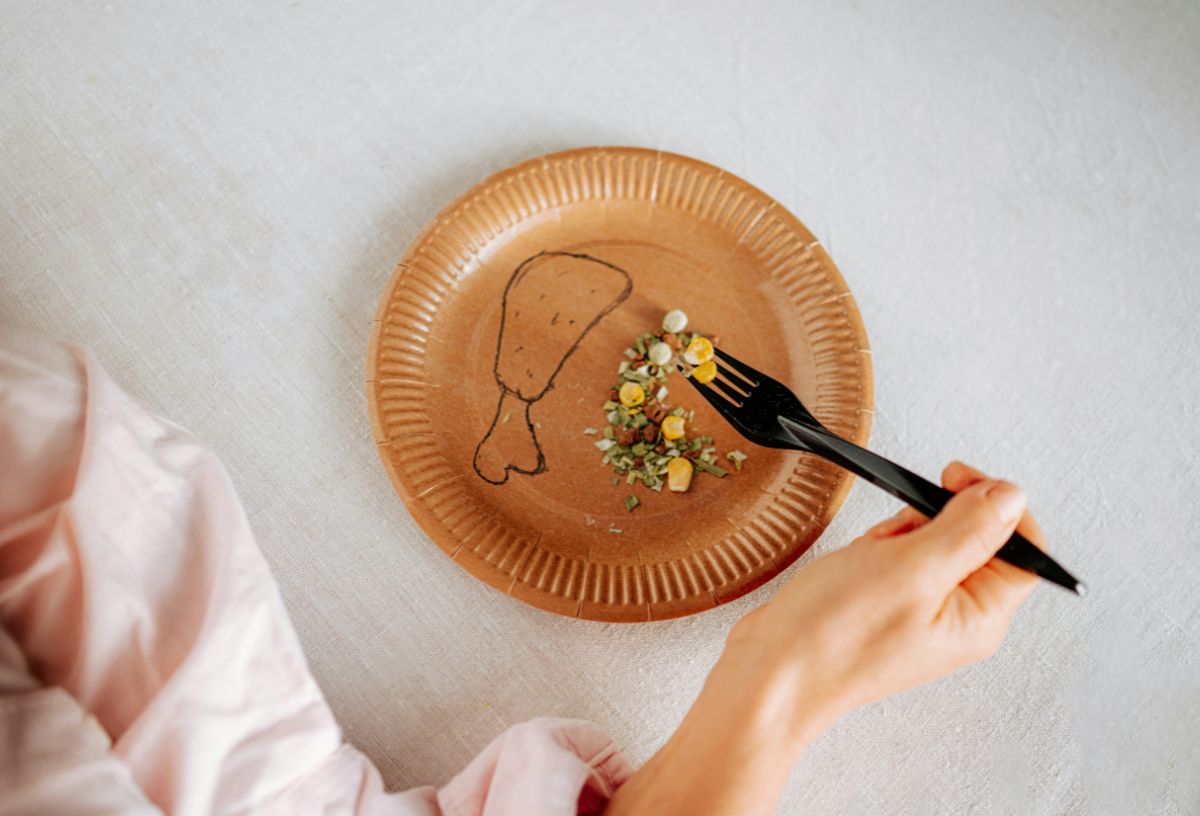 Felülnézetből barna papírtányér, rárajzolt csirkecomb és saláta, fekete villával nő szúr az ételből