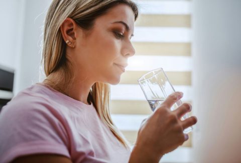 Szőke hajú fiatal nő ablak előtt állva rózsaszín pólóban egy pohár vizet iszik, a fény hátulról világítja meg