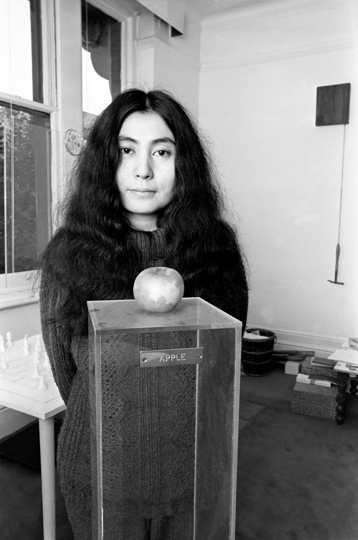Yoko Ono mosolyog egy fekete-fehér képen, előtte egy alma látható