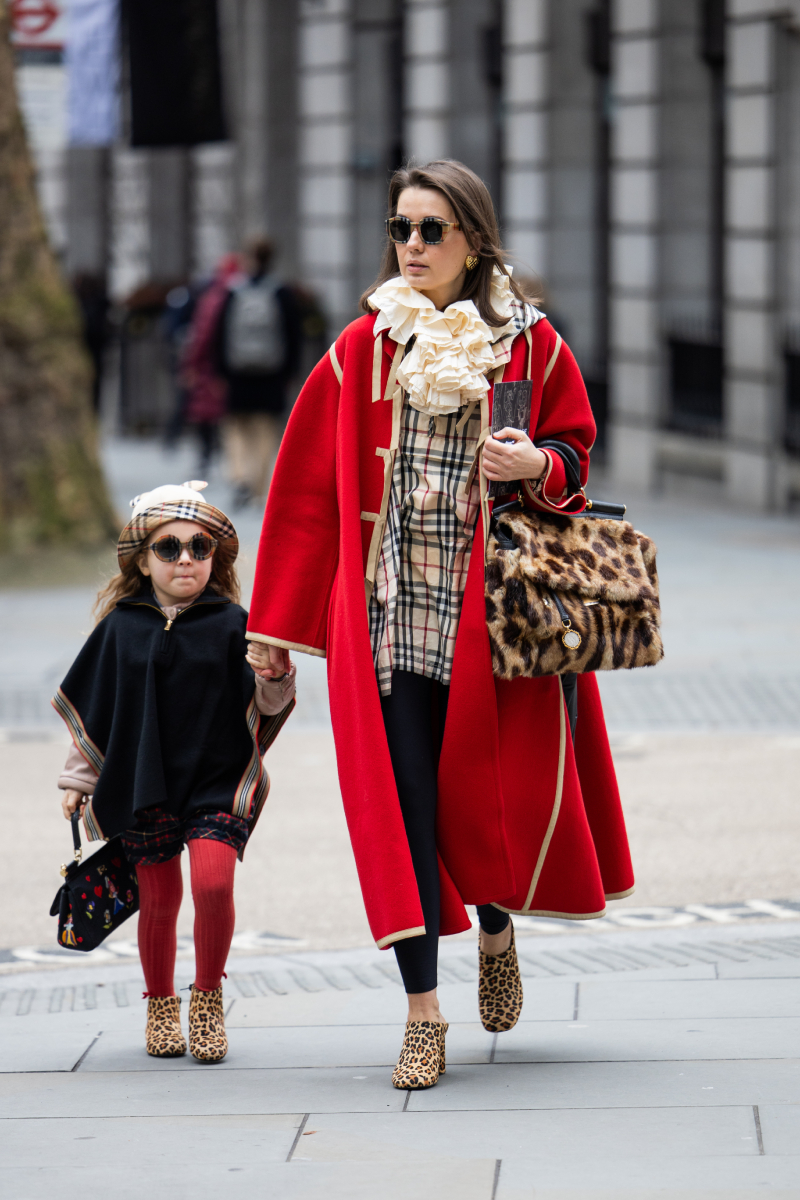 Egy vendég gyermekével együtt, piros oversized kabátban, állatmintás táskával, Burberry dzsekiben és fodros gallérral a Paul Costelloe-bemutató előtt a 2026-os őszi–téli londoni divathéten.