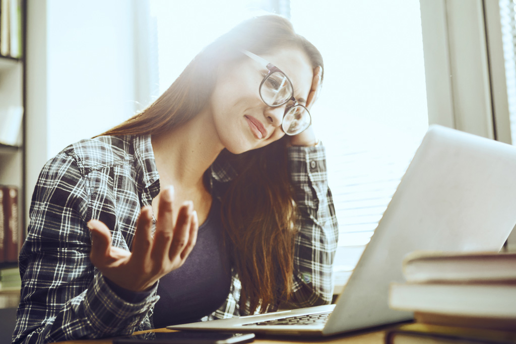 Egy kockás inget viselő szemüveges nő néz értetlenül a laptopja előtt és feltartja az egyik kezét