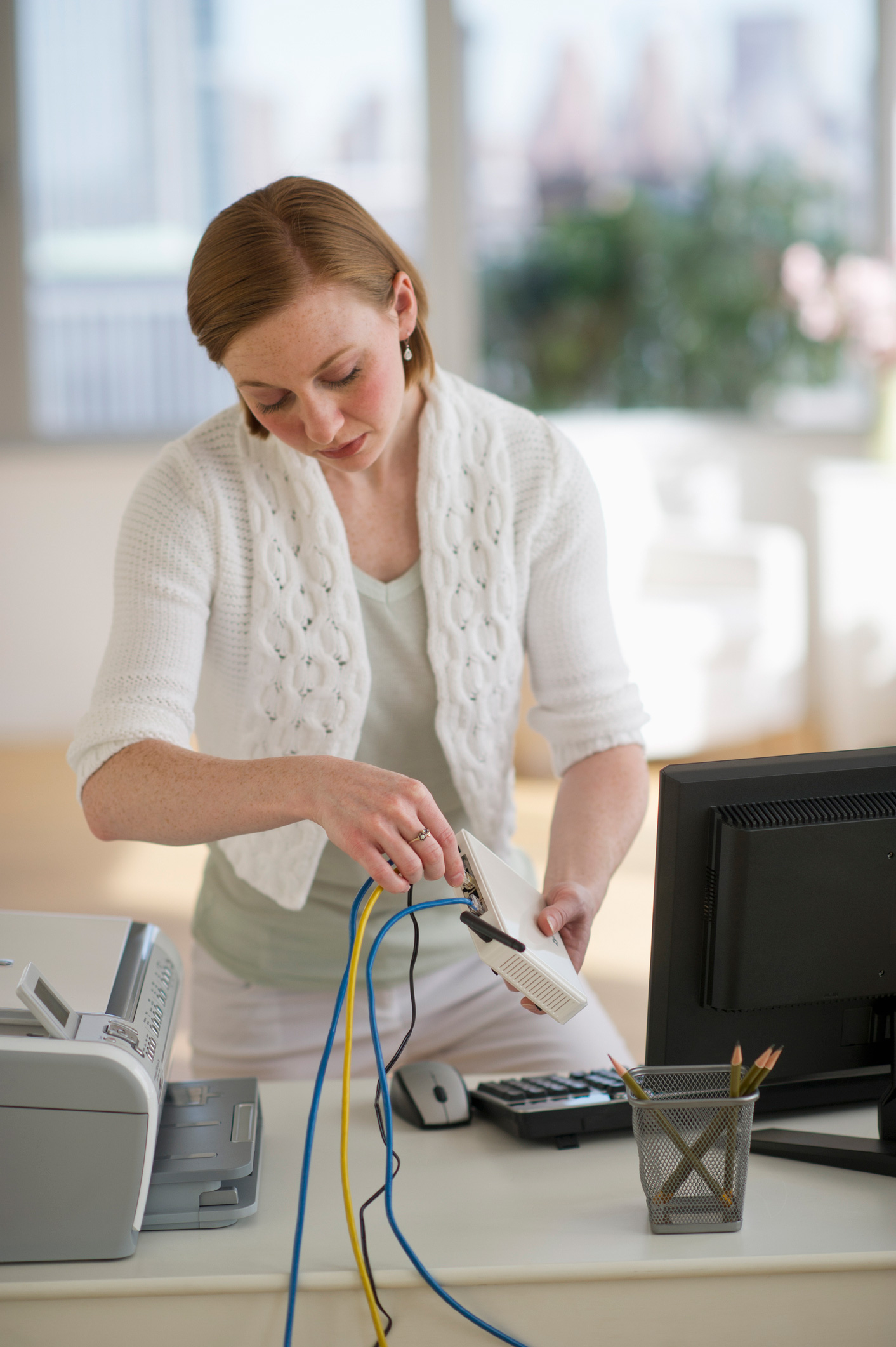 Egy fehér kardigános nő otthon a routerének kábelét ellenőrzi a számítógép mellett