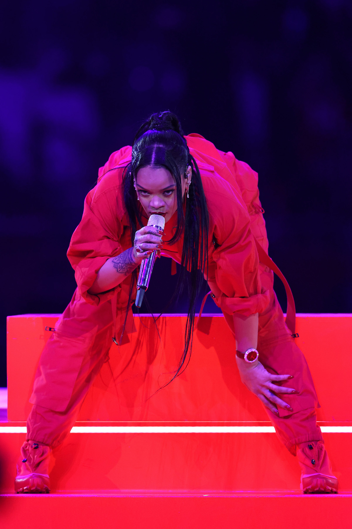Rihanna talpig pirosban, piros háttér előtt előre hajolva mikrofonba énekel