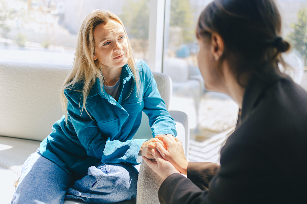 Két középkorú nő egymás kezét fogja a támogatás és az empátia jeleként. Irodai környezet, kollégák, közös problémamegoldás.