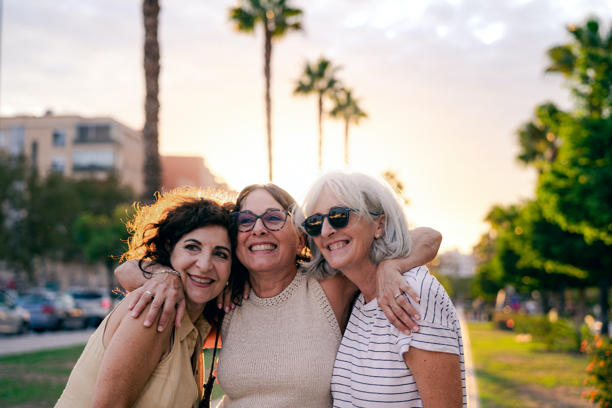 Három életre szóló barát ölelkezik és boldogan mosolyog az utcán naplementekor, ellenfényben. A mögöttük ragyogó napfény körvonalazza az alakjukat, kiemelve az örömüket és a köztük lévő mély baráti köteléket.