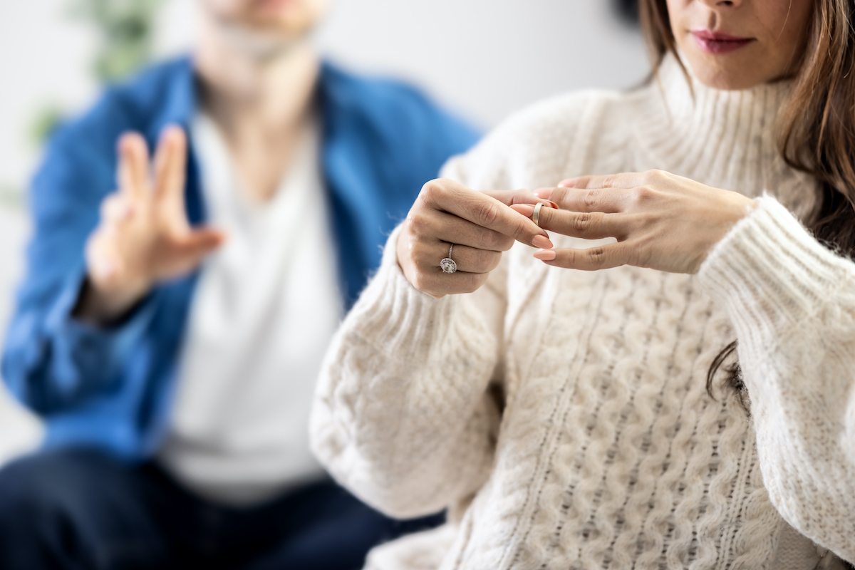 fehér pulóveres nő lehúzza az ujjáról a gyűrűt, a háttérben kék farmeringes férfi