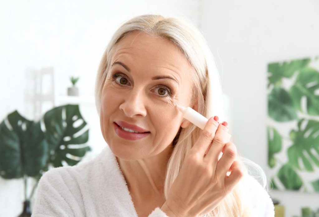 aranyerkenocs-szem-ala | Marie Claire Szőke, hosszú hajú ötvenes éveiben járó nő fehér tubusból krémet nyom a szeme alá szem alatti karikák ellen