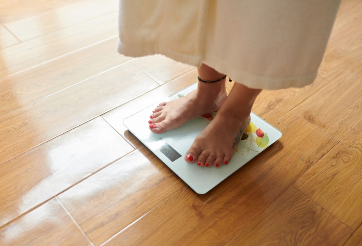 Nő fehér fürdőköntösben fürdőszobai mérlegen áll parkettás szobában mezítláb, egyik bokáján bokakarkötővel