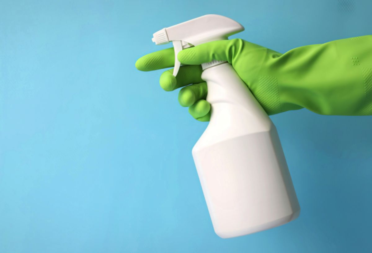 Kék háttér, fehér szórófejes flakon ablakmosó folyadéknak, amit zöld gumikesztyűs kéz tart