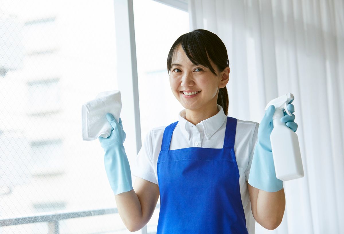 Ázsiai nő kék kötényben, gumikesztyűben hidrogén-peroxidos ablakmosó oldatot tart fehér szórófejes flakonban az egyik kezében, másik kezében fehér törlőruha