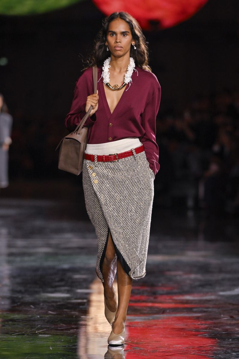 2026-tavasz-trend-library-chic-sikkes-konyvtaros | Marie Claire Színes bőrű modell a Chanel 2026. tavaszi–nyári bemutatóján bordó kardigánban, szürke ceruzaszoknyában, nyakában feltűnő nyaklánc, vállán táska