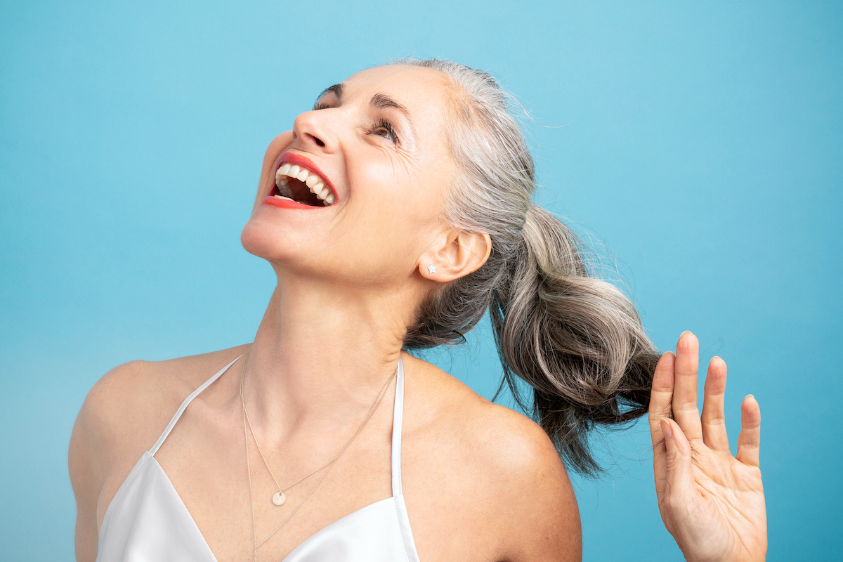 kék háttér előtt hosszú, ősz hajú nő a lófarkához ér és nevet