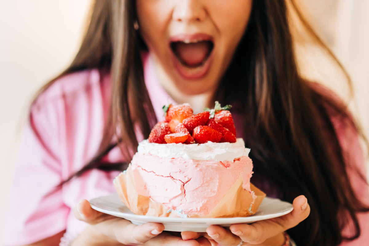 Hosszú, barna hajú nő tátja a száját, egy nagy tál epres sütemény megevésére készül