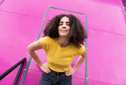 Fiatal, göndör barna hajú nő mosolyogva hajol a kamera fölé sárga felsőben rózsaszín háttér előtt