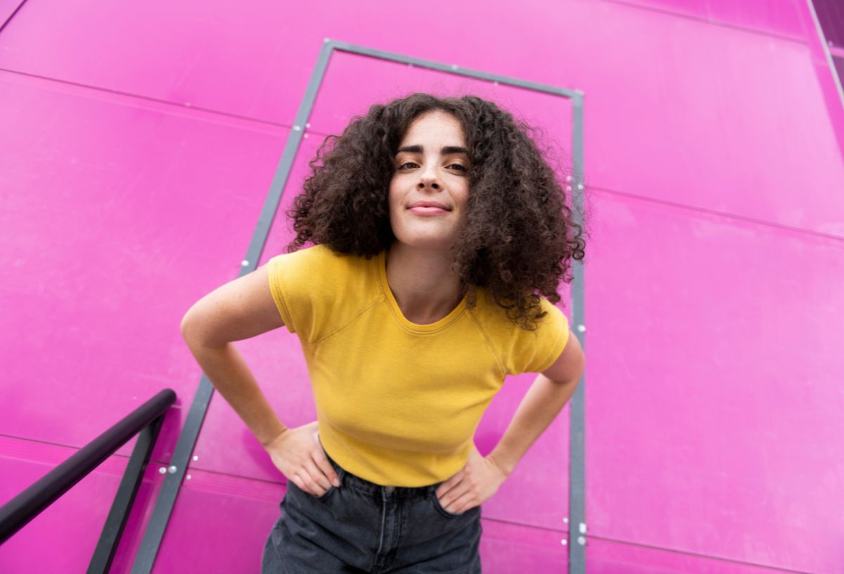 Fiatal, göndör barna hajú nő mosolyogva hajol a kamera fölé sárga felsőben rózsaszín háttér előtt