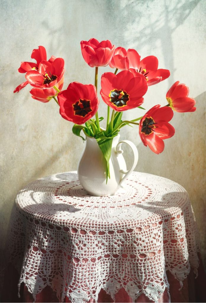Körasztal fehér csipketerítővel letakarva, rajta fehér kerámiakancsóban piros tulipánok