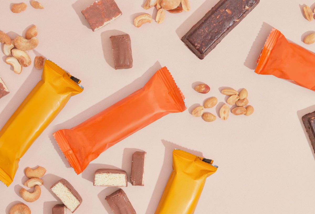 Bézs háttéren narancs és sárga csomagolású fehérjeszeletek, mandula, kesudió