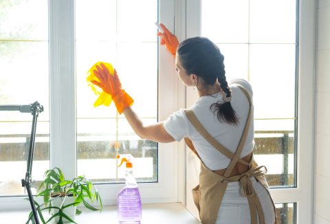 Sötét hajú nő befont hajjal, fehér pólóban és barna kötényben hátulnézetben ablak előtt áll és narancsszínű gumikesztyűben sárga törlőruhával mossa az ablakot, hidrogén-peroxidos ablakpucoló oldat lila szórófejes flakonban mellette a pulton
