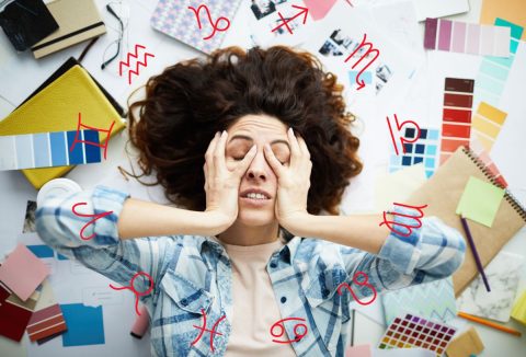 Egy barna hajú nő fogja a fejét, ki van borulva, a nagy rendetlenségben. Horoszkóp szimbólummal.