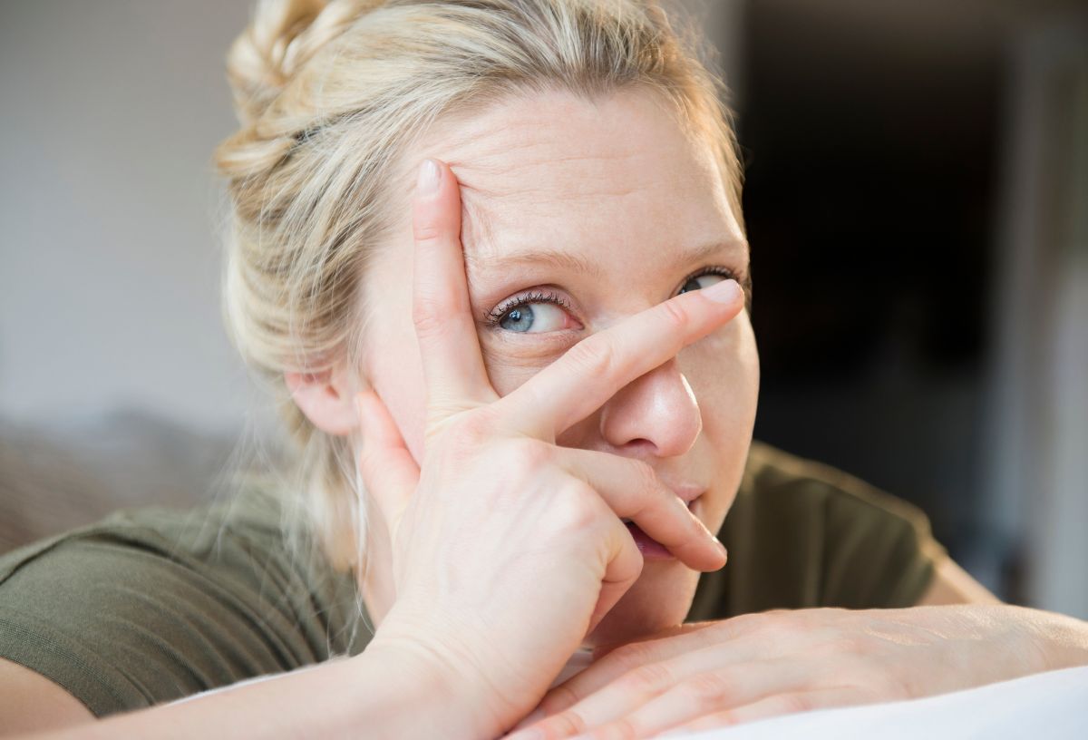 Introvertált szőke hajú, kék szemű nő a kezét az arcához támasztja a közeli fotón és az ujjai között néz ki