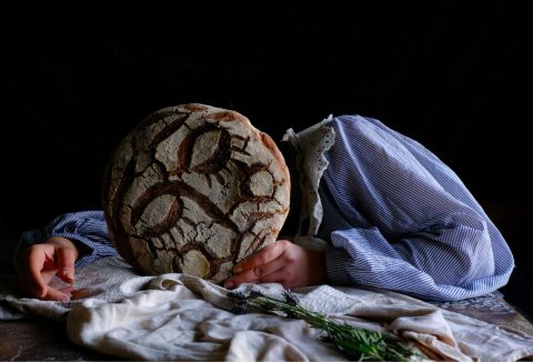 Nő csíkos, buggyos ujjú blúzban, asztalra dől, fejét egy kerek, szomorú arccal díszített kovászos kenyér takarja, fekete háttér