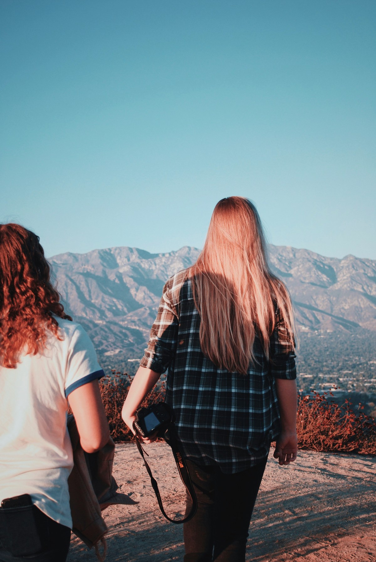 Két nő hegyen kirándul, háttal, a szőke, hosszú hajú nő kezében fényképezőgépet tart.