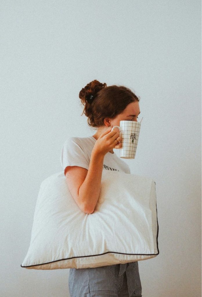 Nő ébredés után, hóna alatt párnával, kezében egy csésze meleg vízzel