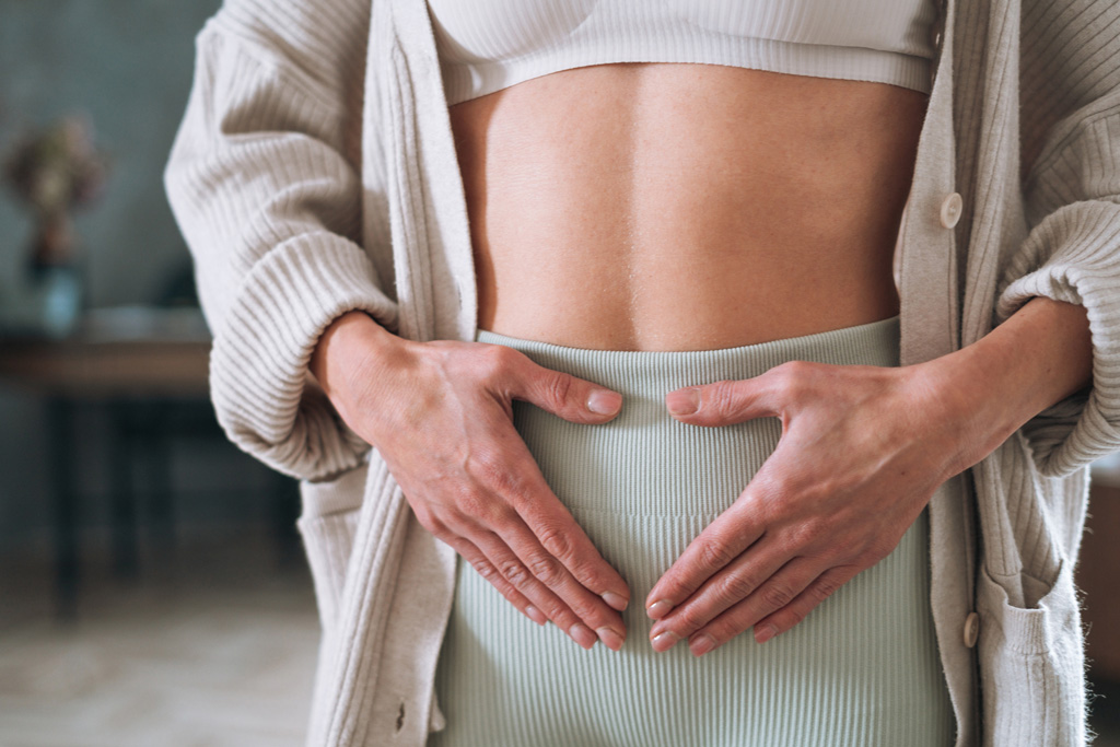 Egy nő középső testrésze látható, fitnesz ruhát visel és a hasát fogja két kezével