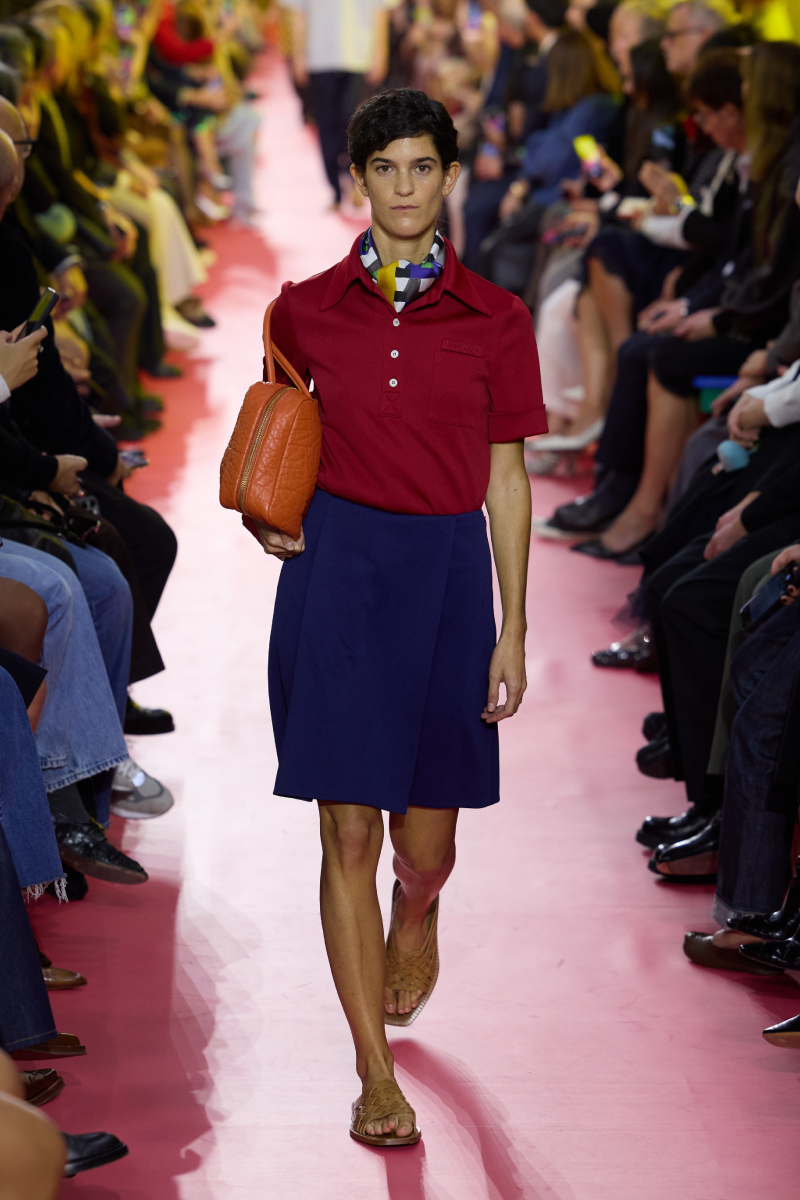 2026-tavasz-trend-library-chic-sikkes-konyvtaros | Marie Claire Rövid hajú modell a Miu Miu 2026. tavaszi–nyári bemutatóján bordó galléros pólóban, kék szoknyában, nyakában kendő, hóna alatt bőrtáska
