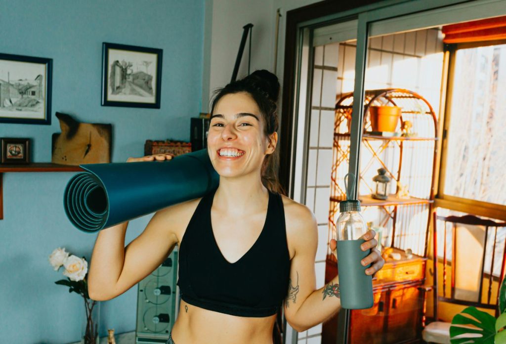 mozgas-lapos-has | Marie Claire Fiatal, sportos nő jógamatraccal a vállán és kulaccsal a kezében boldogan indul edzeni fekete sportmelltartóban az otthonában