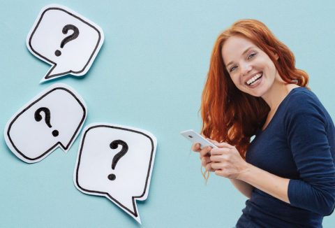 Nő vidáman néz a kamerába telefonozás közben, világoskék háttér, kérdőjelek gondolatbuborékban