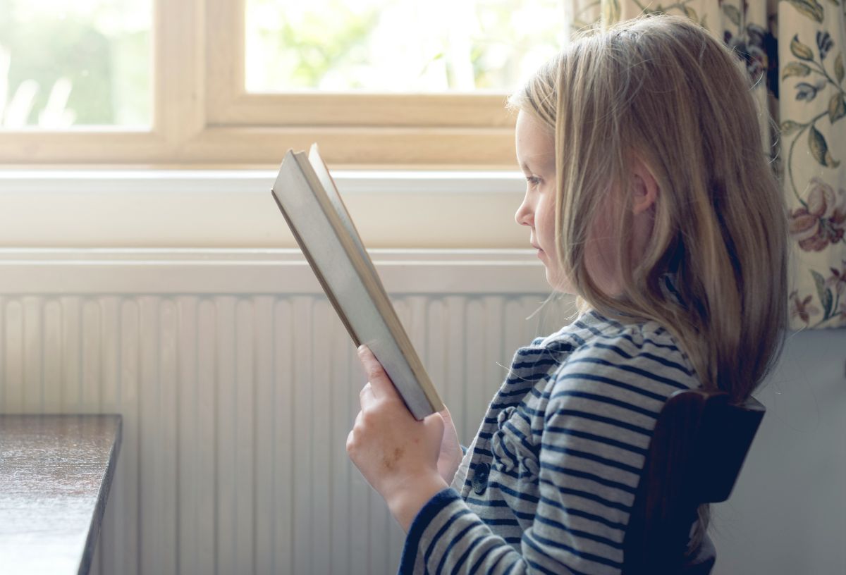 Kislány oldalról könyvvel a kezében ablaknál olvas