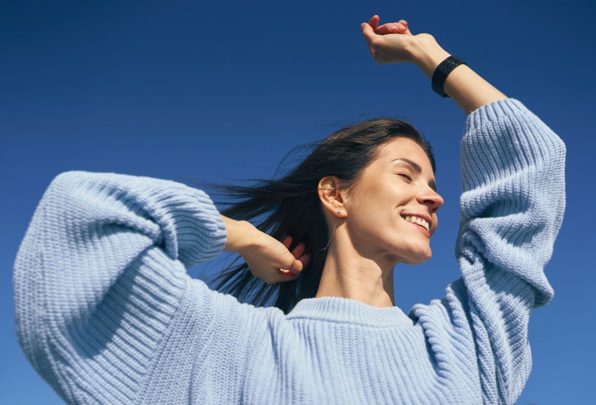 Szép fiatal nő kék égbolt előtt kék pulóverben boldogan emeli magasba a kezét