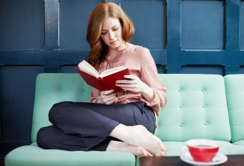 Nő mentazöld kanapén könyvet olvas felhúzott lábakkal kék háttér előtt, az asztalon piros csésze