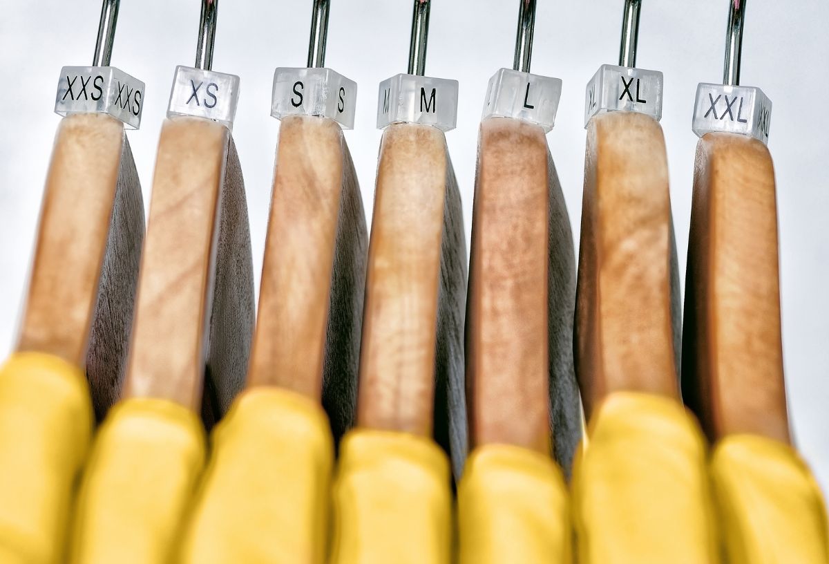 Sárga felsők fa vállfákon, a vállfák nyakán a méretezést jelző műanyag kocka XXS-től XXL-es méretekig