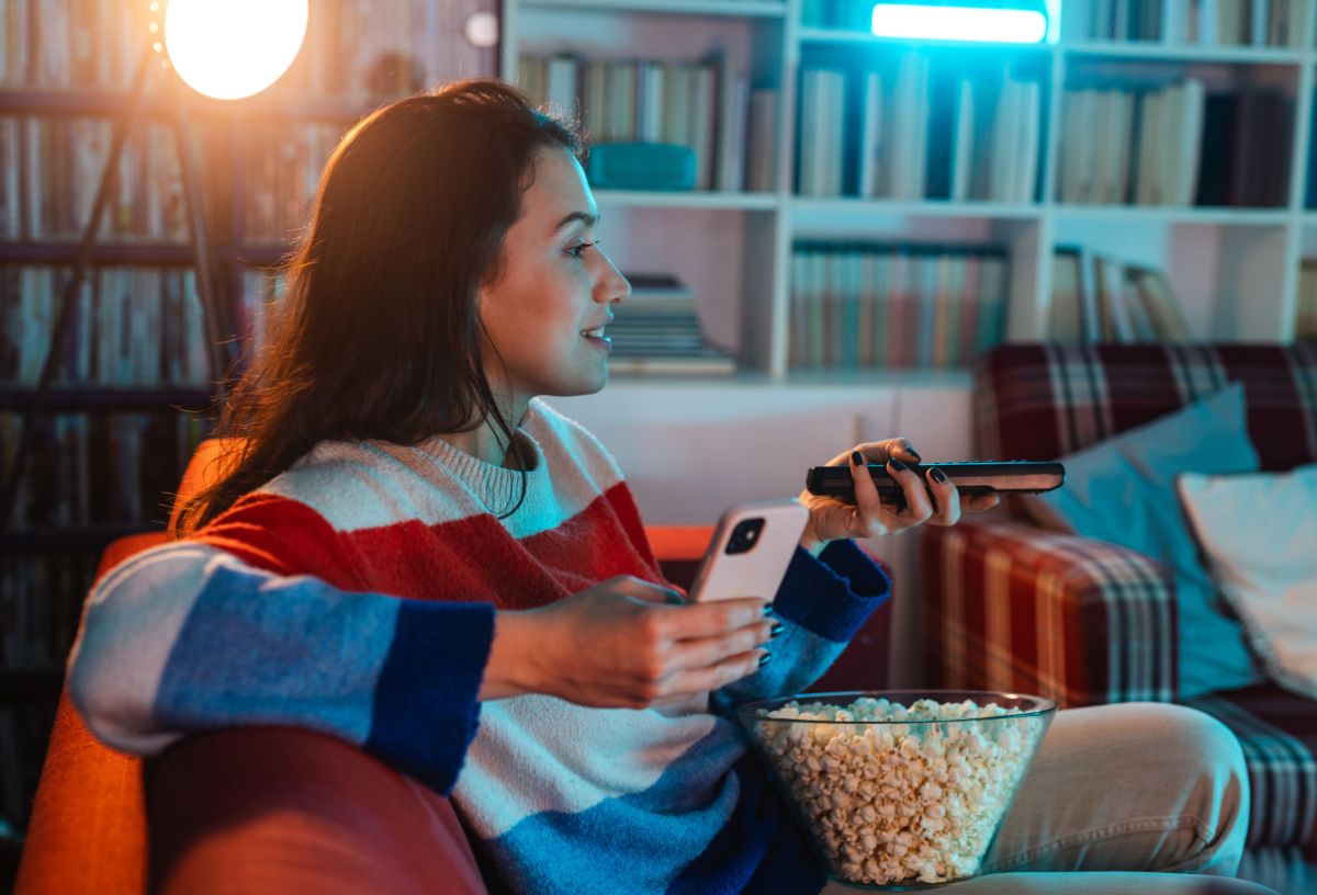 Nő csíkos pulcsiban kanapén könyökölve sorozatot néz, előtte popcorn, egyik kezében távirányító, másikban okostelefon