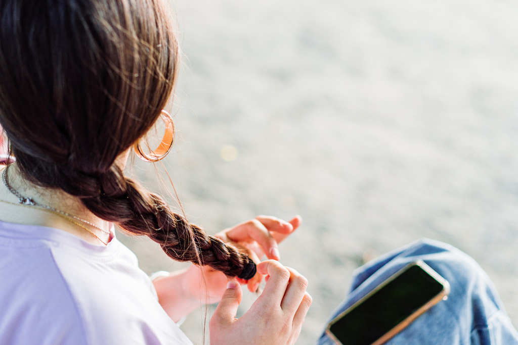 Egy nő a szabadban fonja a haját, és a mobilja a lábán van