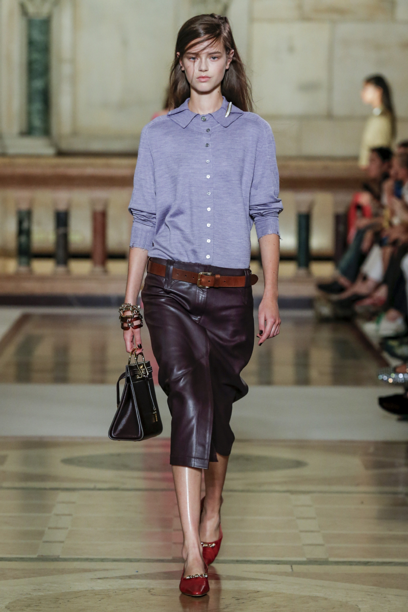 2026-tavasz-trend-library-chic-sikkes-konyvtaros | Marie Claire Barna hajú modell a Tory Burch 2026. tavaszi–nyári bemutatóján kék ingben, fekete bőr ceruzaszoknyában, kézitáskával a kezében