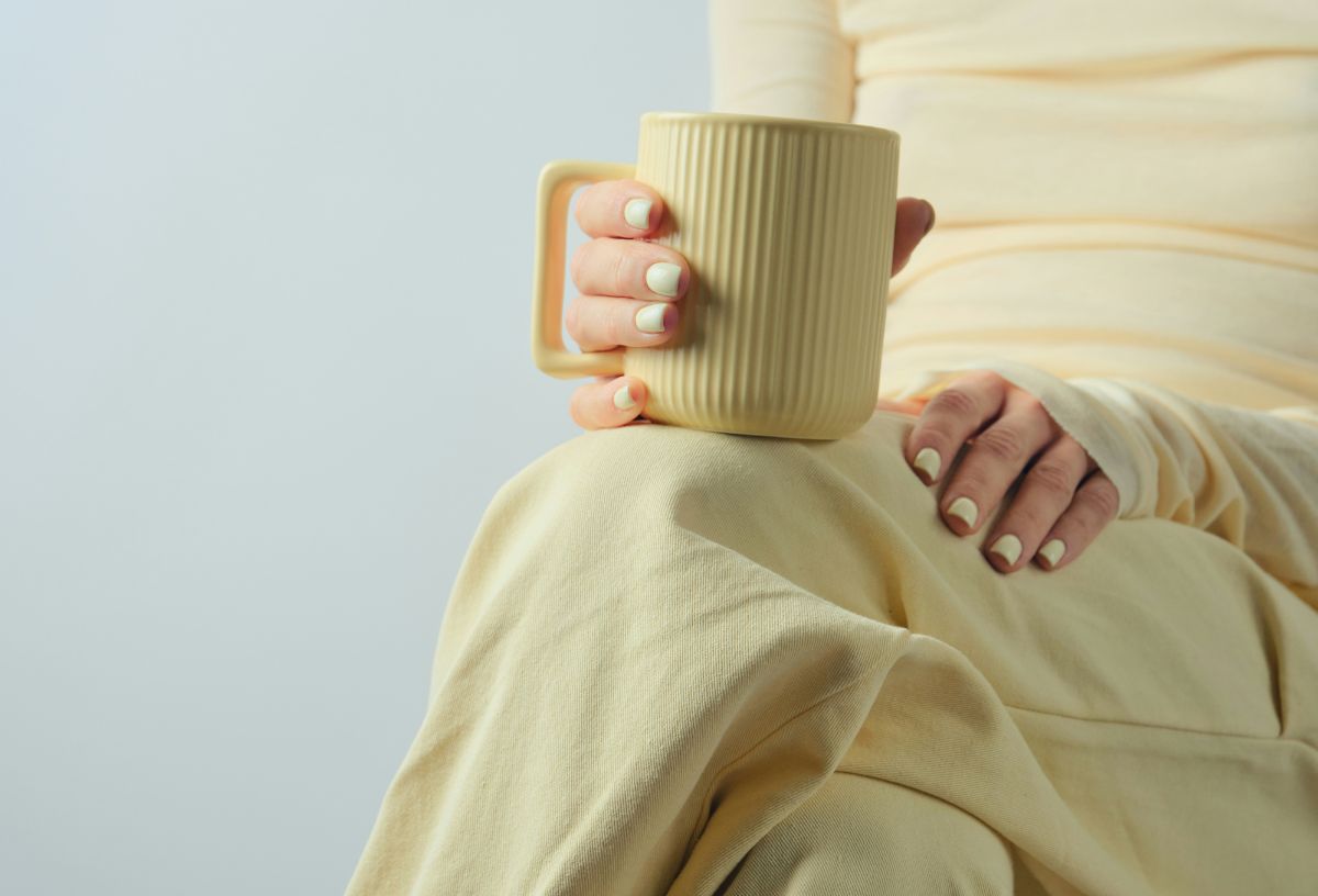 Nő vajsárga körmökkel vajsárga ruhában vajsárga bögrét fog keresztbetett lábbal ülve