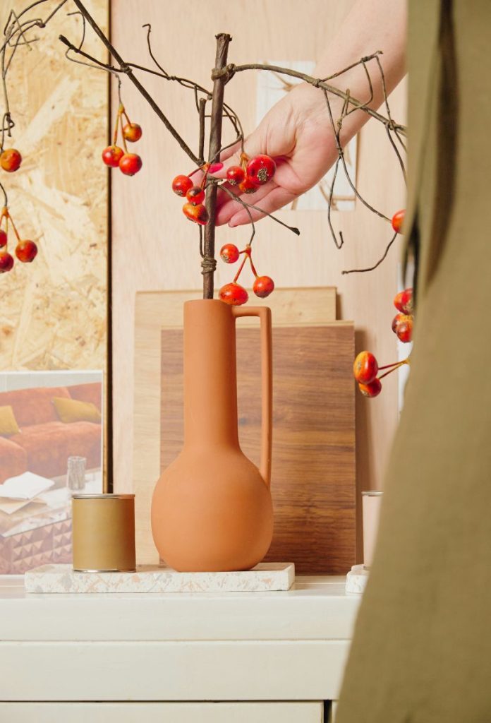 vajsarga-lakberendezes-2026 | Marie Claire Meleg neutrális tónusok vázán, piros bogyós növényt megérintő női kéz, fa elemek a háttérben