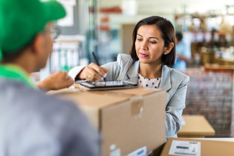 középkorú nő zöld sapkás futártól vesz át nagyméretű csomagot